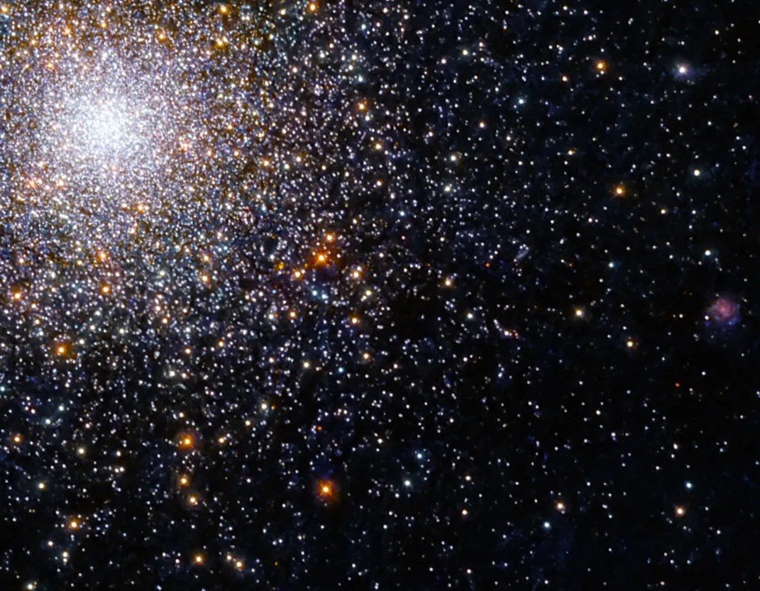 Bright-white spherical cluster of stars in the upper left corner. Stars taper off down and out to the right of the cluster against a black background. Reddish-orange stars dot the scene.