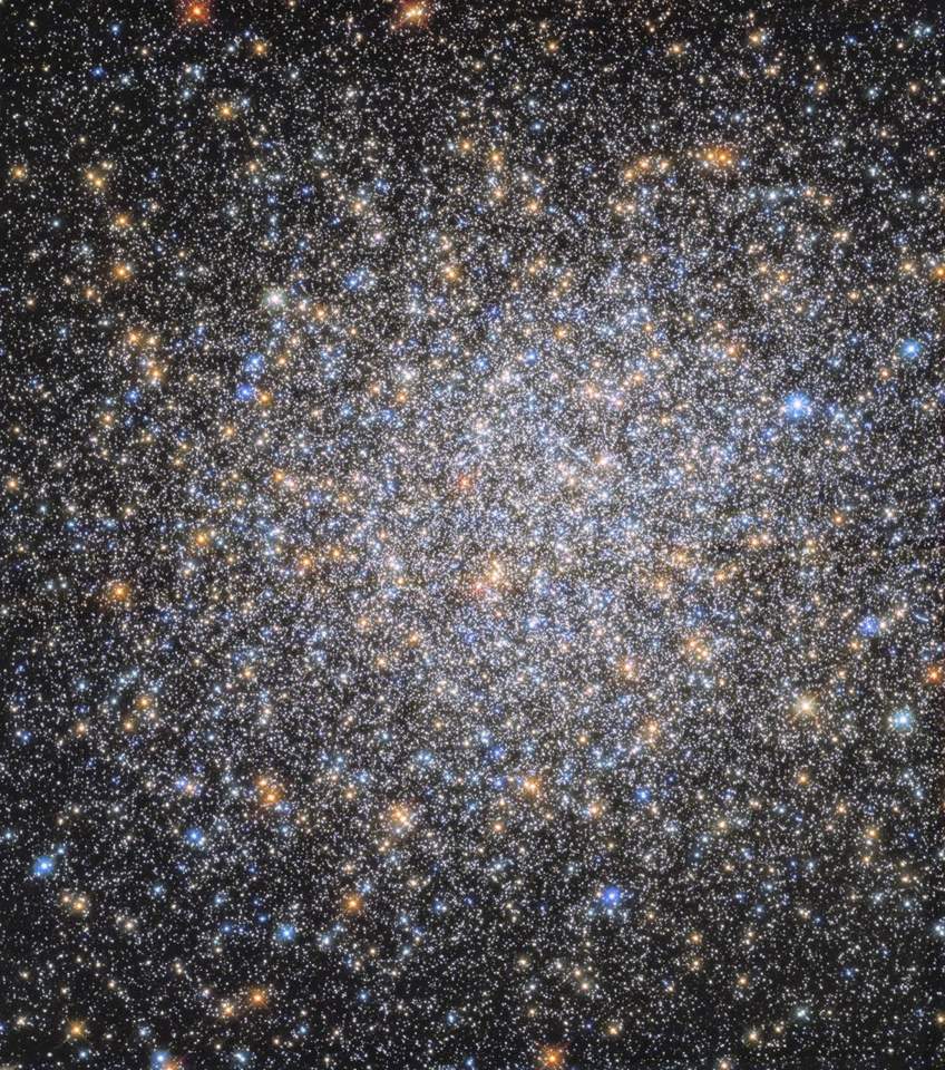 The field is filled with orange, red, yellow, blue, and white stars. They appear as a spherical, dense mass that tapers out toward the edges of the image on a black background.