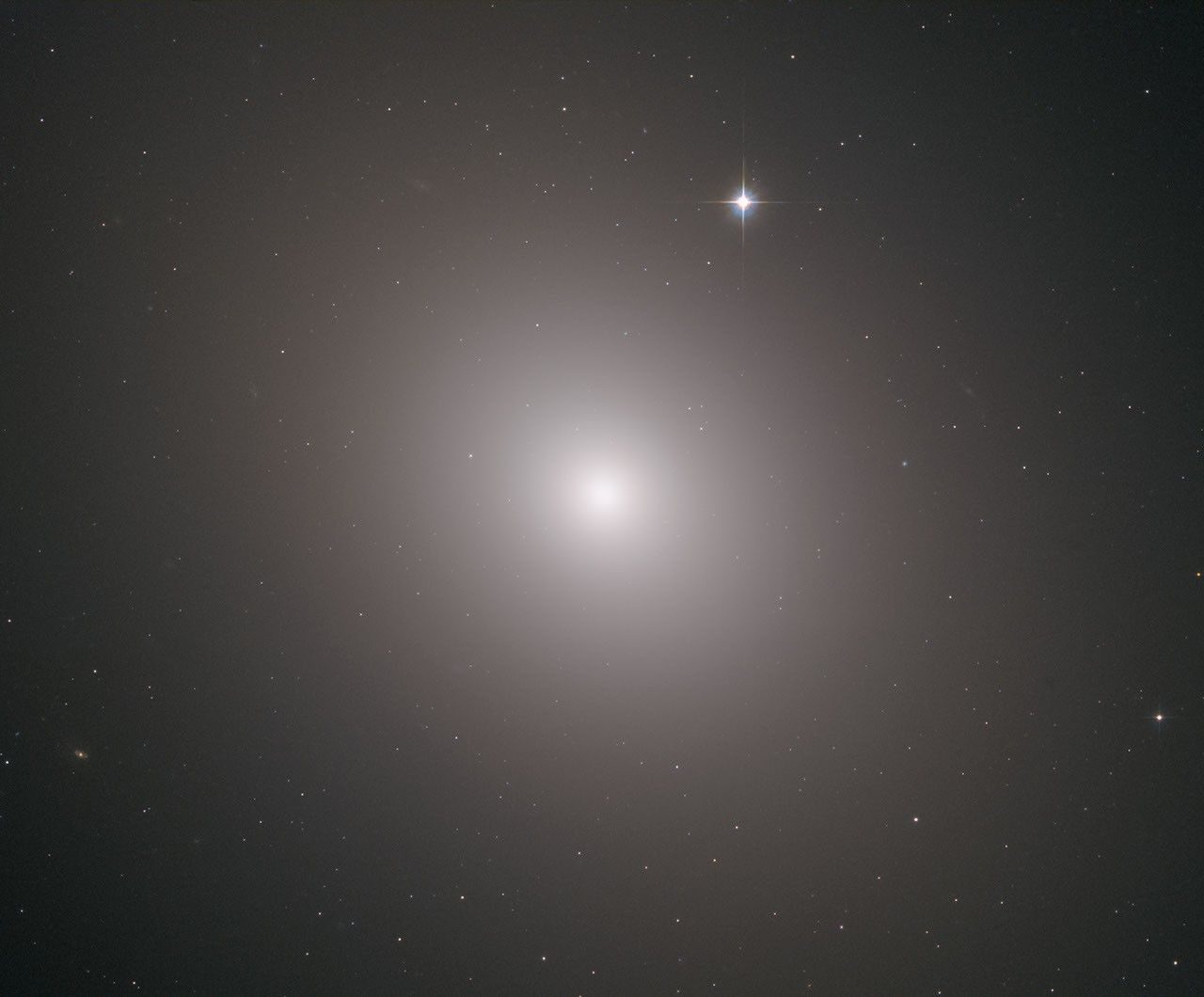 A bright sphere at image center becomes more diffuse and elliptical moving outward from the sphere. One bright foreground stars shines just in the upper-left quadrant of the image. Background stars shine through the galaxy.